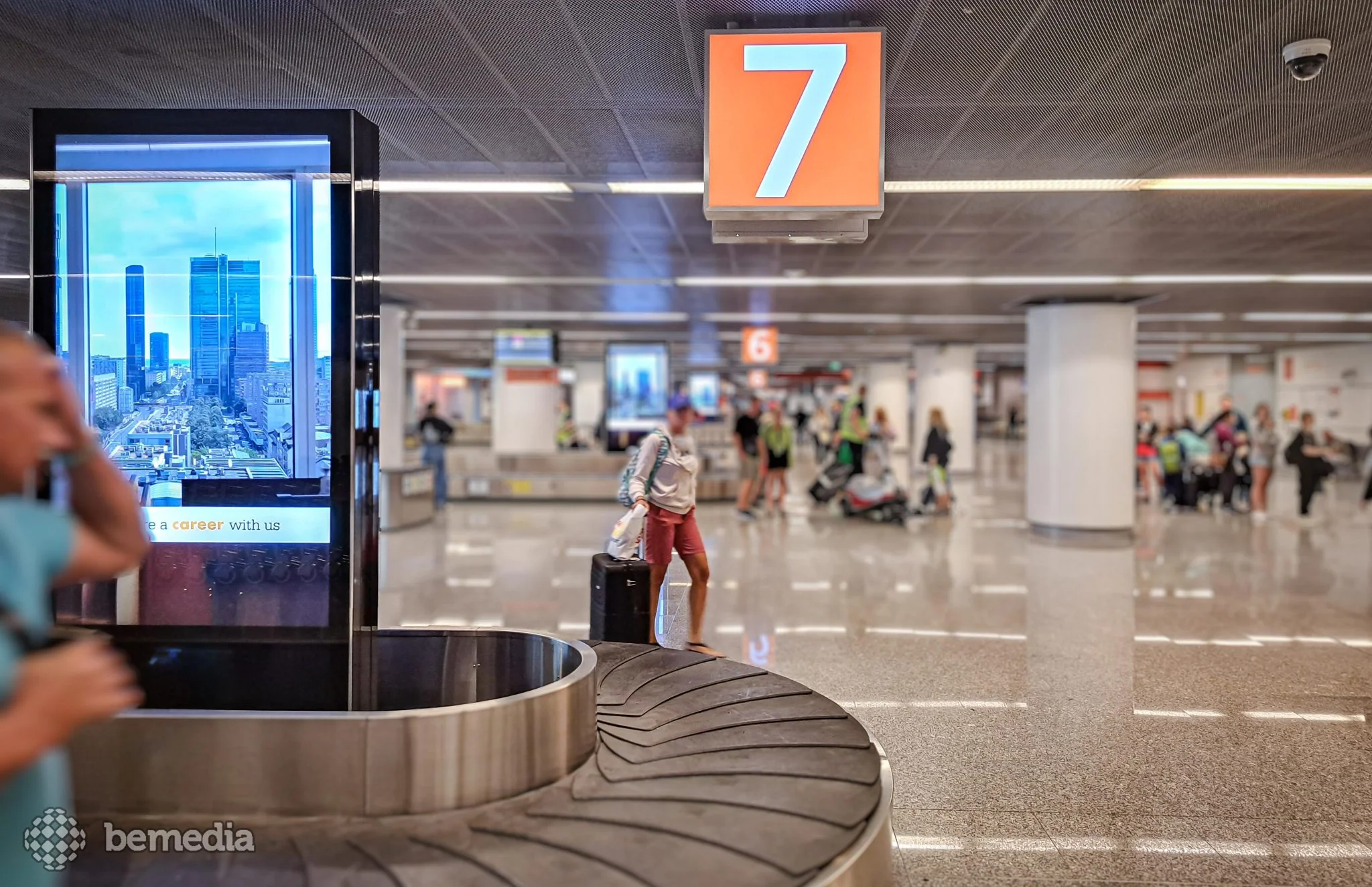 Digital advertising screen beside a baggage carousel in an airport arrivals hall.