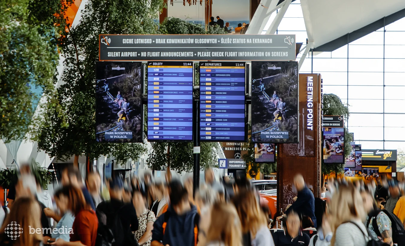 Airport departure information screens surrounded by digital advertising displays in a crowded terminal.