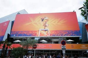 Cannes Film Festival billboard showing a camera operator on a high platform against an orange sunset and crowd of spectators, festival building with banners and logos visible