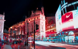 Night view of Piccadilly Circus in London with historic buildings illuminated in red, large LED screens with Nike advertisements, and light trails from passing vehicles captured with long exposure
