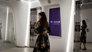 Woman in floral dress standing in front of a Qatar Airways digital display screen showing purple branding with "Going Places Together" slogan in a futuristic white interior space with curved walls and mirrors