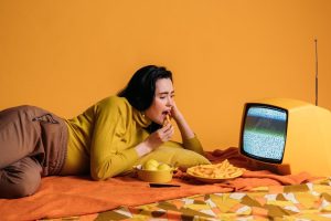 Woman in yellow clothing lying on an orange bed eating snacks while watching a vintage television displaying static, surrounded by chips and fruit in a monochromatic orange-themed setting