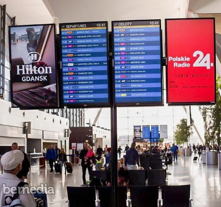Four large digital screens inside Gdańsk Airport displaying flight departure information alongside Hilton Gdańsk and Polskie Radio 24 advertisements, with travelers walking through the terminal.