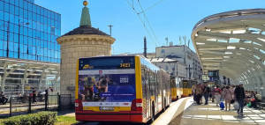 Tył żółto-czerwonego przegubowego autobusu MZA w Warszawie z reklamą Igrzysk Paraolimpijskich Paryż 2024, stojącego na przystanku w słoneczny dzień.