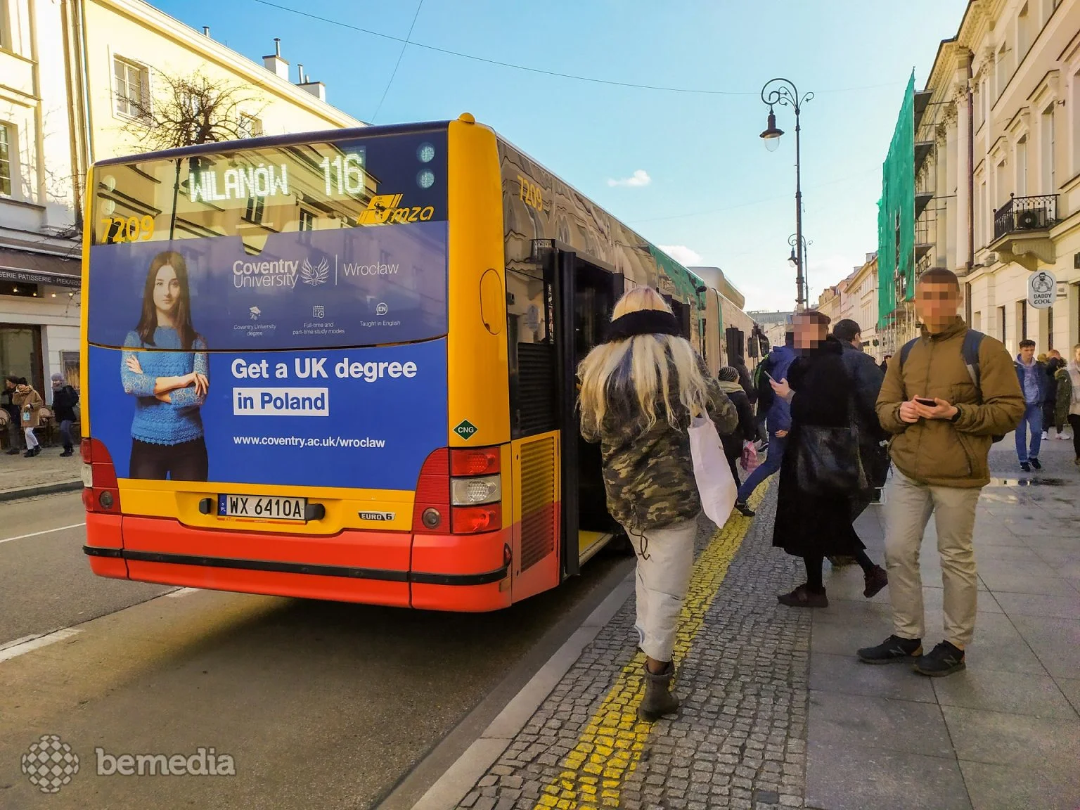 Żółto-czerwony autobus miejski linii 116 jadący w kierunku Wilanowa, z reklamą uczelni Coventry University Wrocław na tylnej szybie, zatrzymany na przystanku przy miejskim chodniku.