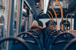 Widok z wnętrza piętrowego autobusu ukazujący rzędy niebieskich siedzeń, charakterystyczne żółte poręcze oraz pasażerów siedzących tyłem do obiektywu, z wyraźnym napisem Piccadilly Circus na elektronicznej tablicy informacyjnej.