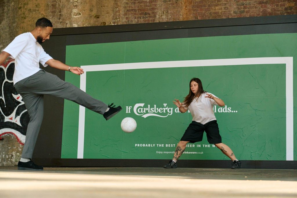 Dwie osoby grające w piłkę nożną na ulicy przed dużym billboardem reklamowym Carlsberga z zielonym tłem