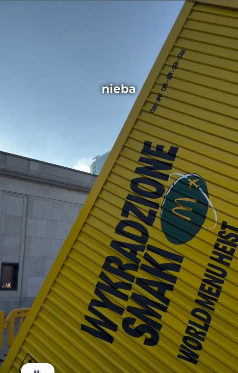 Żółty, falisty kontener reklamowy McDonald's z dużym czarnym napisem "WYKRADZIONE SMAKI" oraz logo marki w zielonym owalu. Obok widoczny jest angielski napis "WORLD MENU HEIST". W tle fragment budynku i błękitne niebo z naklejoną naklejką "nieba".