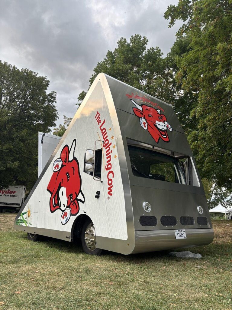 Srebrny samochód dostawczy marki The Laughing Cow o unikalnym, trójkątnym kształcie przypominającym plaster serka (tzw. 'Wedgemobile'), zaparkowany na trawie wśród drzew. Pojazd ozdobiony jest charakterystycznym logo czerwonej, uśmiechniętej krowy oraz napisem 'The Laughing Cow'.