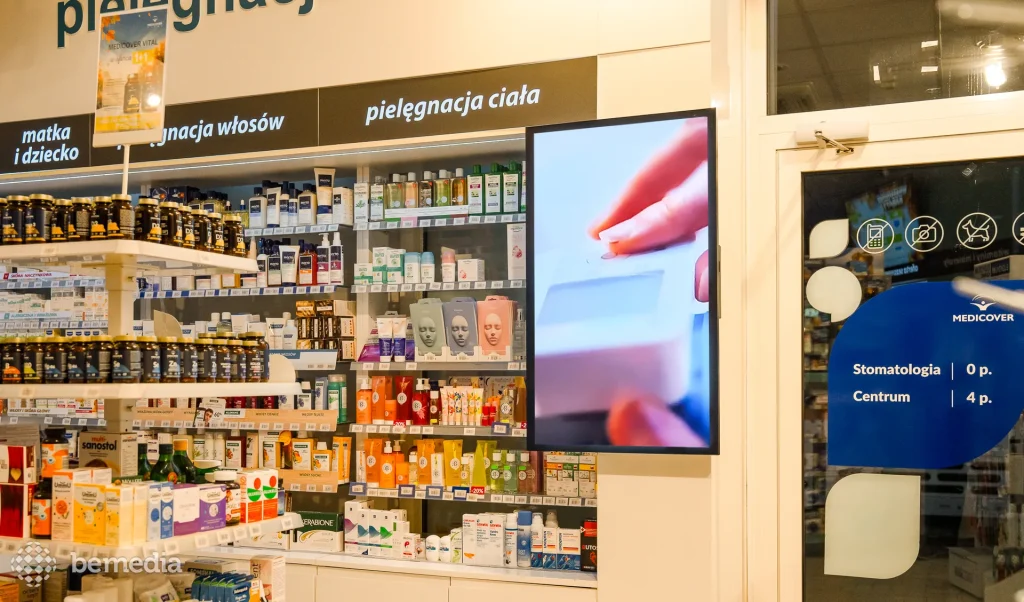 Pionowy ekran LCD z reklamą w aptece, obok regałów z kosmetykami i suplementami